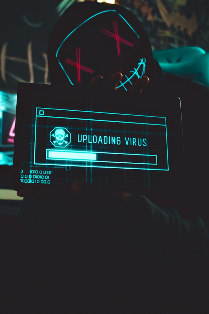 why-choose-us Hacker in a dark room holding a tablet with a virus uploading screen, representing cyber threat.